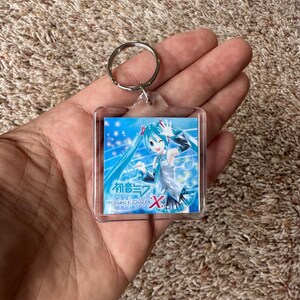 May include: A square clear acrylic keychain featuring an anime character with long teal hair and a white outfit. The insert displays the text Hatsune Miku Project DIVA X with a blue background, sparkling light effects, and musical notes.