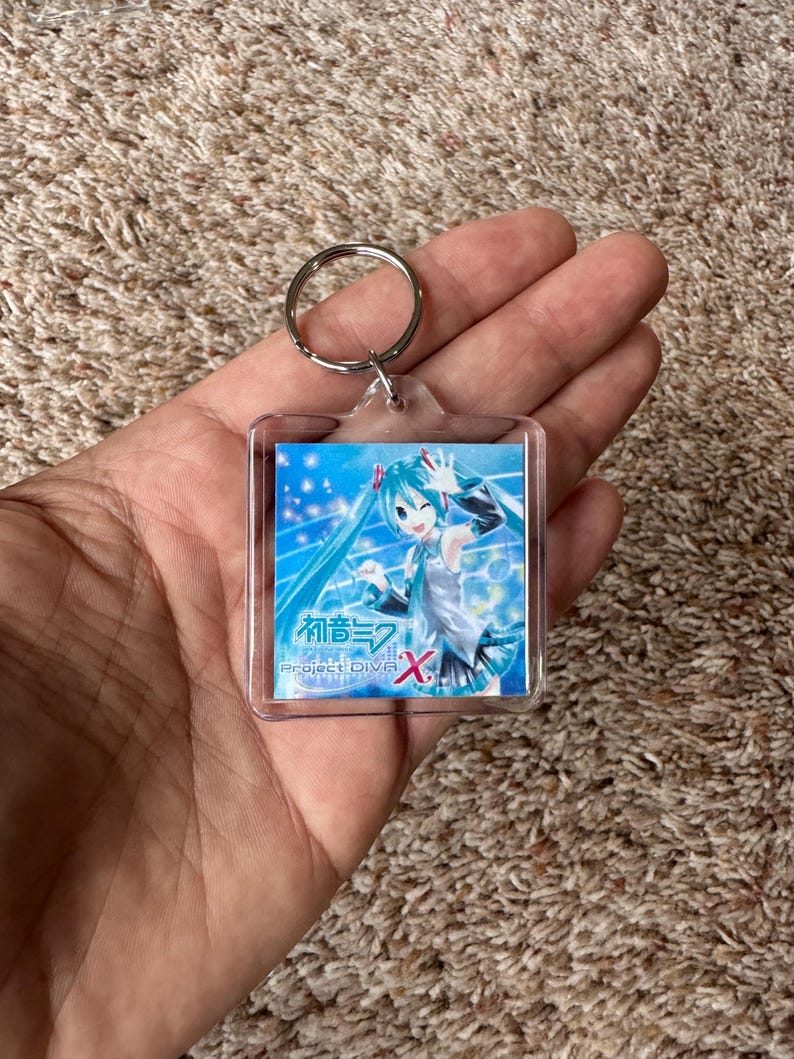May include: A square clear acrylic keychain featuring an anime character with long teal hair and a white outfit. The insert displays the text Hatsune Miku Project DIVA X with a blue background, sparkling light effects, and musical notes.