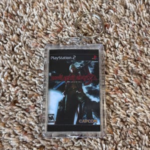 May include: A clear plastic keychain featuring a PlayStation 2 game cover for Devil May Cry 3: Dante's Awakening. The game cover art shows a character with a sword. The keychain has a silver metal ring.