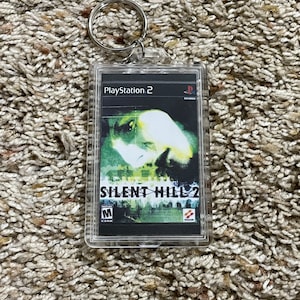 May include: A clear acrylic keychain featuring the cover art of the PlayStation 2 game "Silent Hill 2." The artwork displays a blurred, green-toned image with the game's title in white. A silver key ring is attached at the top.