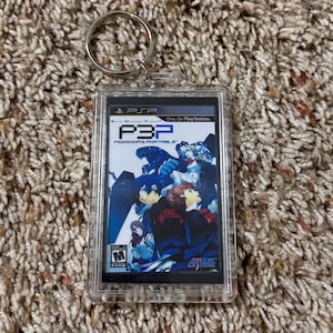 May include: A rectangular keychain featuring the cover art of the video game "Persona 3 Portable" for the PSP. The artwork depicts several characters in a blue and red color scheme, with the game's title prominently displayed. The keychain has a silver key ring.