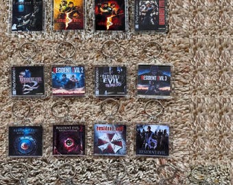 May include: A collection of seventeen square acrylic keychains, each featuring a miniature graphic print of a Resident Evil video game cover art. Each keychain includes a silver-toned metal split ring attached to the top for hanging.