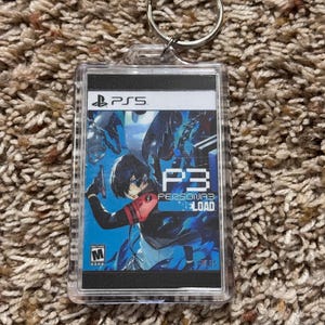 May include: A clear acrylic keychain featuring the PS5 logo and the game title "P3 Persona 3 Reload." The keychain has a silver keyring and displays a colorful game graphic with a character in a blue and black outfit.
