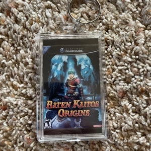 May include: A clear acrylic keychain featuring the Baten Kaitos Origins video game cover art. The artwork depicts a character in red and brown armor with a blue and white background. The keychain includes a silver metal keyring.