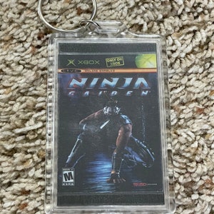 May include: Clear rectangular keychain featuring the cover art of the video game "Ninja Gaiden" for Xbox. The artwork depicts a ninja in a dark setting. The keychain has a silver metal ring.