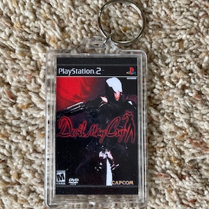 May include: A clear acrylic keychain featuring the cover art from the video game "Devil May Cry" for PlayStation 2. The artwork depicts a character with a gun against a red and black background. The keychain includes a silver metal ring.