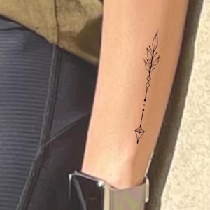 May include: A black, minimalist arrow tattoo design on the forearm. The tattoo features a leaf, diamond, and arrow-shaped elements. The person is wearing a silver watch with a black screen and a light-coloured band.