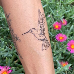 May include: A black and white hummingbird tattoo on an arm. The hummingbird is shown with its wings extended. The background includes green foliage and bright pink flowers with yellow centres.