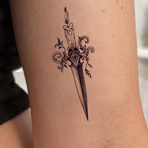 May include: A black and white tattoo design featuring a dagger with a candle on top. The candle has a lit flame, and the dagger is decorated with ornate floral and scrollwork details. The design is a tattoo.