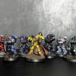 May include: A collection of miniature figurines of Space Marines, each painted in different colors, including red, blue, purple, yellow, black, white, and green. The figures are posed in action stances and stand on detailed bases.