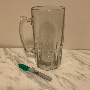 May include: A clear glass beer mug with a handle. The mug is sitting on a white marble surface. A green and gray Sharpie fine point marker is lying next to the mug.