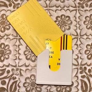 Puede incluir: Un tarjetero blanco con una tarjeta amarilla parcialmente insertada. La tarjeta amarilla tiene las palabras "TACO CAT GOAT CHEESE PIZZA" impresas. El tarjetero está sobre una superficie estampada.