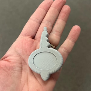 May include: A light grey, 3D-printed pendant with an oval centre and a decorative top. The pendant is held in a hand, showcasing its shape and design. The pendant is approximately 5 cm long.