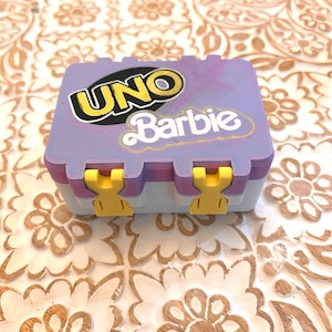 May include: A purple and white UNO Barbie game box with yellow clasps. The box features the UNO logo and the Barbie logo in white. The box is on a wooden surface with a floral pattern.