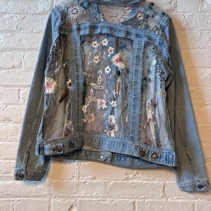 May include: A light blue denim jacket featuring sheer mesh panels on the back and sides. The mesh sections are decorated with intricate, multi-colored floral embroidery in shades of pink, white, and blue, with long-sleeved denim arms and a collar.