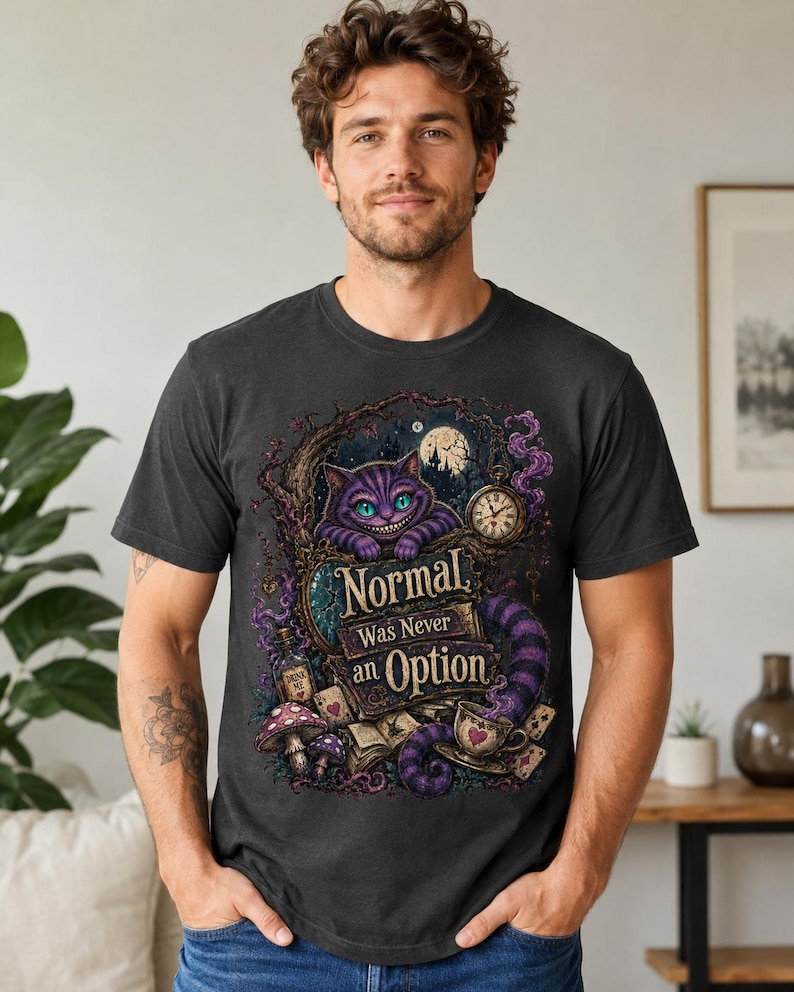 May include: A charcoal gray t-shirt featuring a graphic of a purple striped cat with a wide grin, a pocket watch, a teacup, mushrooms, and playing cards. The text reads, "Normal Was Never an Option," set against a dark, whimsical forest background.