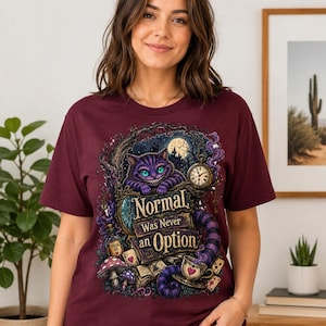 May include: A maroon short-sleeved t-shirt featuring a graphic print of a purple striped cat, a pocket watch, mushrooms, and playing cards. The text on the shirt reads, "Normal Was Never an Option," surrounded by a dark, whimsical forest scene.