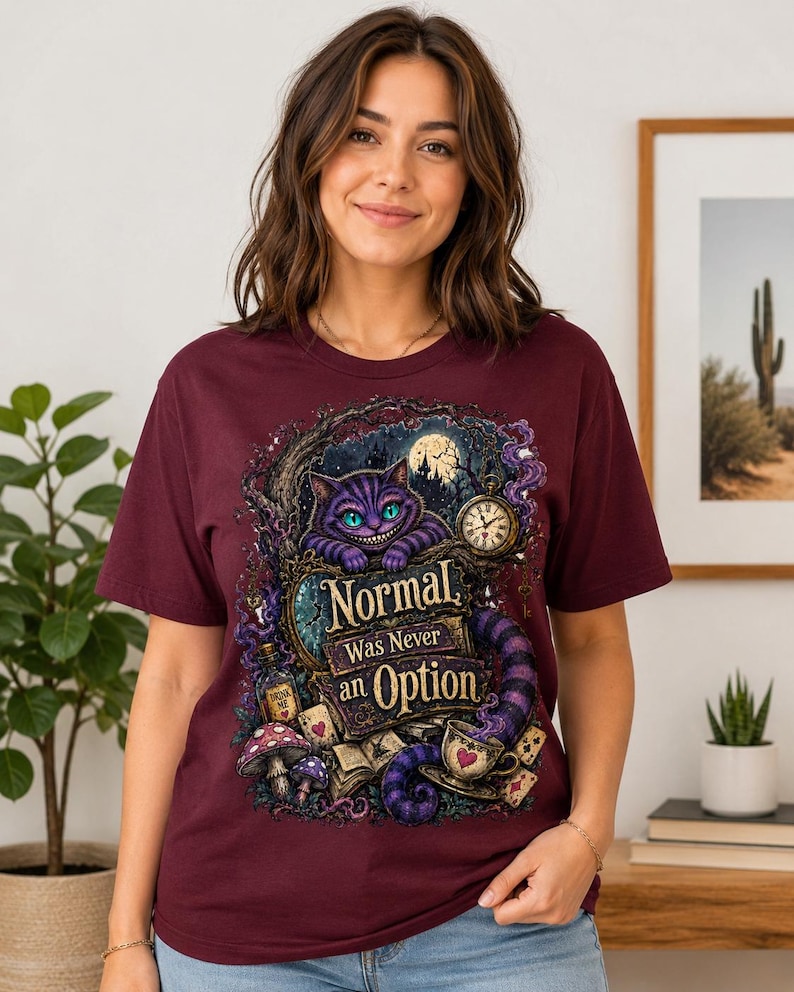 May include: A maroon short-sleeved t-shirt featuring a graphic print of a purple striped cat, a pocket watch, mushrooms, and playing cards. The text on the shirt reads, "Normal Was Never an Option," surrounded by a dark, whimsical forest scene.