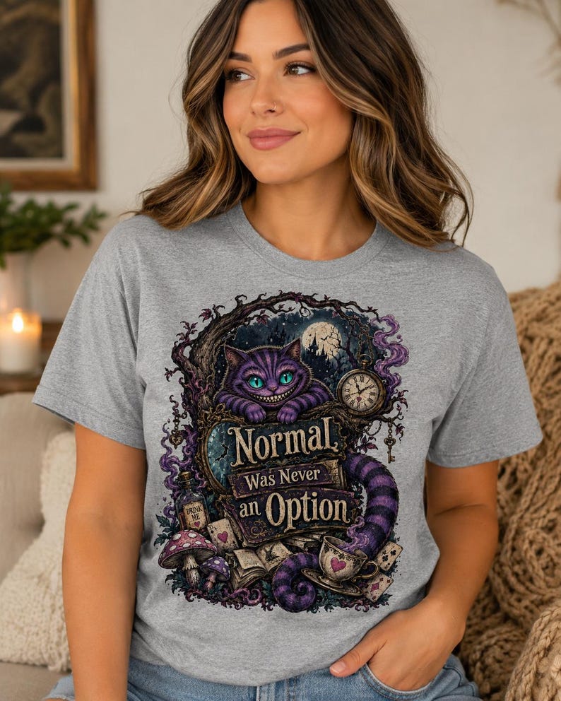 May include: A heather gray t-shirt featuring a graphic print of a purple striped cat with a wide grin, a pocket watch, mushrooms, and playing cards. The text on the shirt reads, Normal Was Never an Option, in a decorative, dark-themed fantasy style.