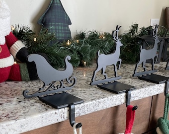 May include: Four black metal stocking holders with a sleigh and reindeer designs. The holders are attached to a wooden countertop with stockings hanging from them.