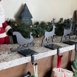 May include: Four black metal stocking holders with a sleigh and reindeer designs. The holders are attached to a wooden countertop with stockings hanging from them.