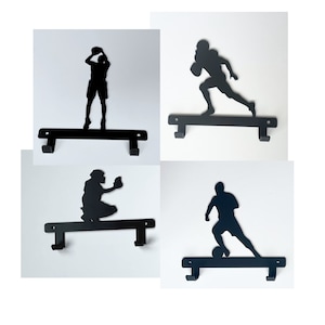 May include: Four black metal sports-themed coat racks. Each rack features a silhouette of a different sport: baseball, football, soccer, and basketball. Each rack has two hooks for hanging items.