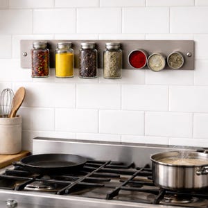 May include: A stainless steel magnetic spice rack with glass jars and metal containers filled with various spices. The rack is mounted on a white tiled wall above a stove with a pan and pot.