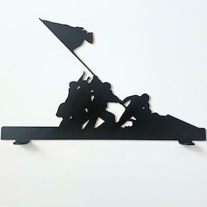 May include: Black metal silhouette wall art depicting a group of soldiers raising a flag on a hill. The soldiers are silhouetted against a white background.