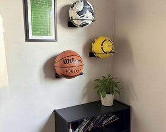 May include: Three sports balls displayed on black wall shelves: a soccer ball, a basketball with "Wilson JORDAN" text, and a yellow ball. A framed soccer field print hangs above the basketball. A small plant in a white pot sits below the shelves.