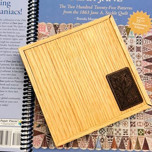 May include: A light-colored wooden tray with a dark rectangular emblem featuring an illustration and the text "FIELD OF SEAMS." The tray is placed on top of a book with spiral binding and text.