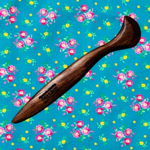 May include: A dark brown wooden tool with a curved handle and pointed end, possibly for crafting or woodworking. The text "FIELD & SEAMS" is visible on the tool. The tool is set against a teal floral patterned background.