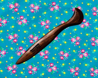 May include: A dark brown wooden tool with a curved handle and pointed end, possibly for crafting or woodworking. The text "FIELD & SEAMS" is visible on the tool. The tool is set against a teal floral patterned background.