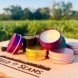 May include: Five small round metal tins with lids. The tins are in various colors, including pink, gold, and purple. The tins are sitting on a wooden surface with the words "FIELD of SEAMS" engraved on it.
