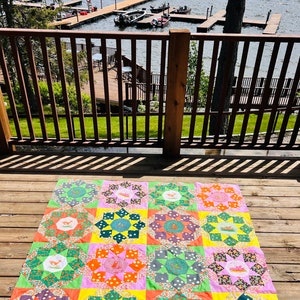 Könnte beinhalten: Ein farbenfroher Patchwork-Quilt mit einem sich wiederholenden Sternmuster. Jedes Quadrat zeigt ein zentrales Medaillon mit einer Fuchsdarstellung, umgeben von leuchtenden Blumen- und Tupfenstoffen. Der Quilt liegt auf einer Holzterrasse mit Blick auf einen See.
