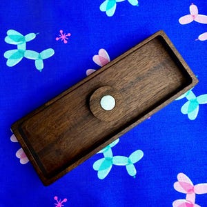 May include: A rectangular dark brown wooden tray with a smooth finish and a centred circular wooden accent piece featuring a small, light-coloured circular inlay. The tray has raised edges and a deep, natural wood grain texture throughout the surface.