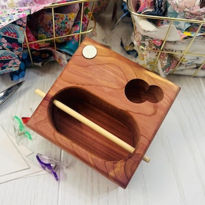 May include: A rectangular cedar wood organizer featuring a large oval cutout with a horizontal wooden dowel across the center, a smaller circular cutout, and a single white circular inlay on the top surface with visible wood grain patterns.