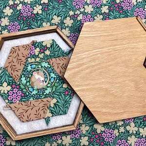May include: Hexagonal wooden box with a light wood lid and a dark wood clasp. Inside, a fabric-lined compartment features a patchwork design with a mouse illustration. The box rests on a floral patterned fabric.