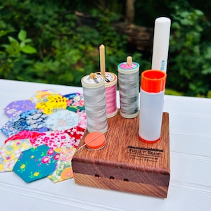 May include: A wooden sewing organiser with three spools of thread, a glue stick, and a needle threader. The spools of thread are pink, grey, and grey. The organiser has the text "FIELD & SEAMS" engraved on the front.