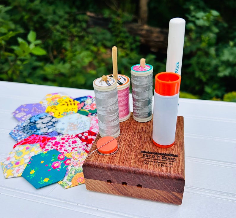 May include: A wooden sewing organizer with three spools of thread, a glue stick, and a needle threader. The spools of thread are pink, gray, and gray. The organizer has the text "FIELD & SEAMS" engraved on the front.