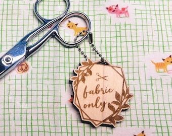 May include: A pair of silver scissors rests on a green and white patterned fabric. A wooden tag with the words "fabric only" is attached to the scissors. The fabric has a repeating pattern of small dogs.