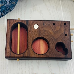 May include: A handcrafted wooden puzzle with a dark brown exterior and reddish-brown interior elements. The puzzle features various circular and oval cutouts, along with wooden dowels and a central sliding piece. The wood grain is visible throughout the design.