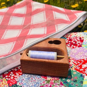 May include: A wooden thread spool holder with a light blue spool of thread, resting on a colorful patchwork quilt. A pink and white checkered bag is in the background, set in a field of yellow flowers.
