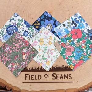 May include: A collection of seven floral patterned fabric squares in various colors, including blue, pink, and green. The squares are arranged on a wooden surface with the text "FIELD OF SEAMS" engraved on it. The fabrics feature different floral designs.