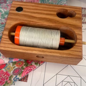 Peut inclure: Un porte-fil en bois avec une bobine de fil blanc et des accents orange. Le support a une forme rectangulaire avec une ouverture incurvée pour le fil et un petit aimant sur le dessus. Du tissu floral est visible en arrière-plan.