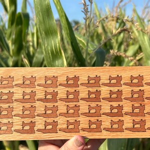 May include: A wooden rectangular sign with a repeating pattern of sewing machine silhouettes. The sign is made of light brown wood with a darker brown wood inlay.
