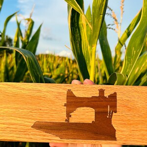 May include: A wooden cutting board with a silhouette of a sewing machine carved into the surface. The board is light brown with a natural wood grain.
