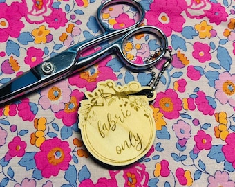 May include: A pair of silver scissors rests on a floral fabric with pink and yellow flowers. A wooden tag with the words "fabric only" is attached to the scissors.