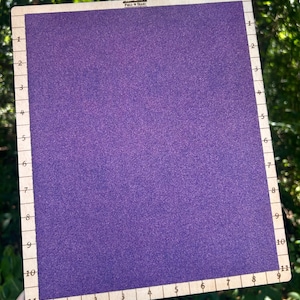 May include: A rectangular purple needlepoint board with a wooden frame. The frame has numbered markings along the edges. The text "FIELD SEAM" is printed at the top. The board is held by a hand.