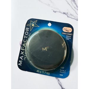 May include: A Max Factor Pan-Cake makeup compact in a dark shade, packaged in a blue and gold blister pack. The compact is round with the letters "MF" in gold. The package indicates the product weighs 1.7 oz.