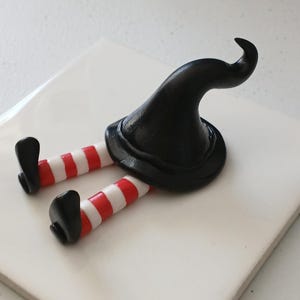 May include: A black witch figurine with red and white striped legs and a pointed black hat. The figurine is lying on its back on a white surface.